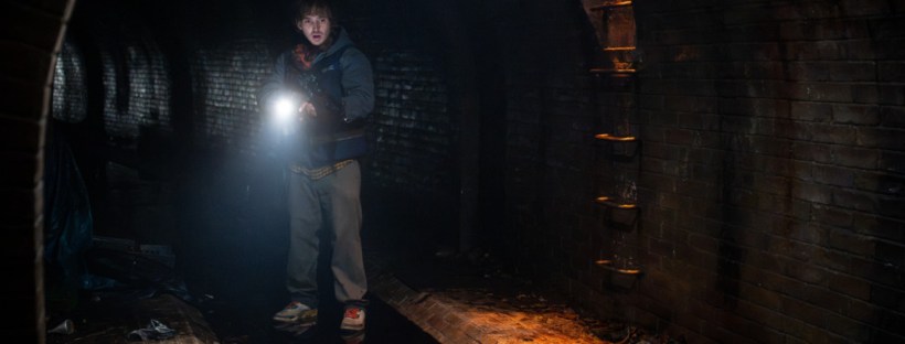 An image of the actor Austin Abrams as the character Bryan in the movie Resident Evil. In the movie, he's standing in a dark sewer tunnel, using the flashlight on his phone.